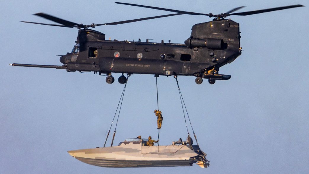 one of an MH-47 Chinook sling loading a stealthy Combat Craft Assault (CCA) boat while one of its crew, most likely a SWCC, fast ropes down into it in mid-air.