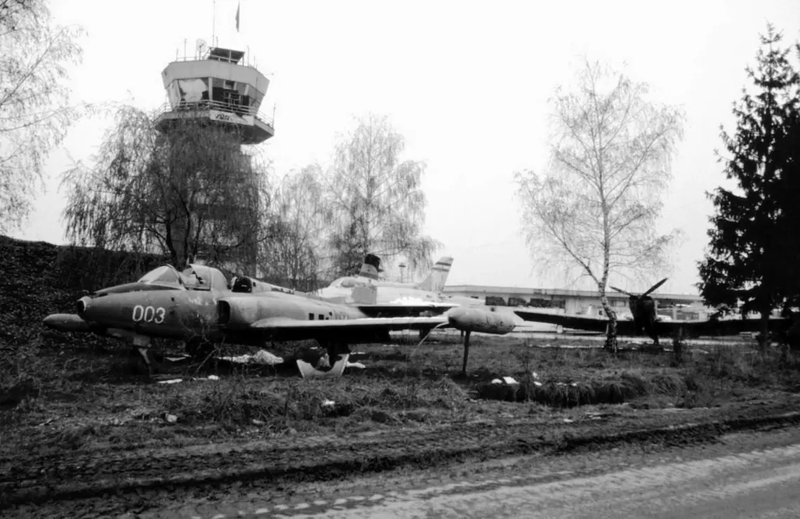A photograph taken by Nick Weight on December 23, 1993, depicting the G-3 aircraft, ev. no. 23003, positioned on the grassy area of Sarajevo's Butmir airport. The aircraft appears abandoned, symbolizing the state of the country it was produced in. In the background, the control tower of the airport is visible.